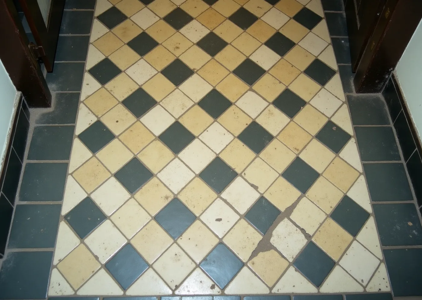 An old entrance-hall floor of cream tiles set on the diagonal with small dark insets, worn and stained where it has been walked.